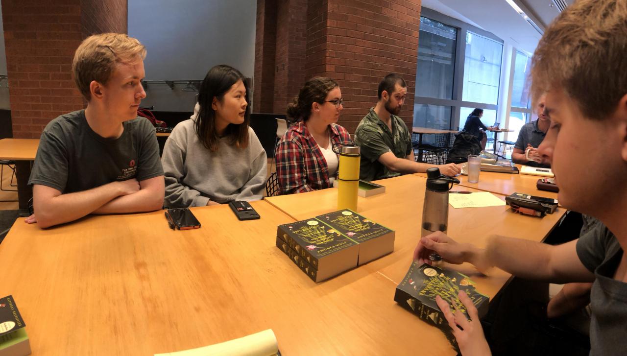 Students at a wooden table, all looking over the same book while having a discussion.