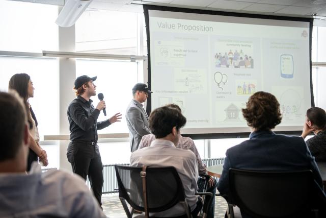Three students at the front of a room presenting from a projector screen to a group of people
