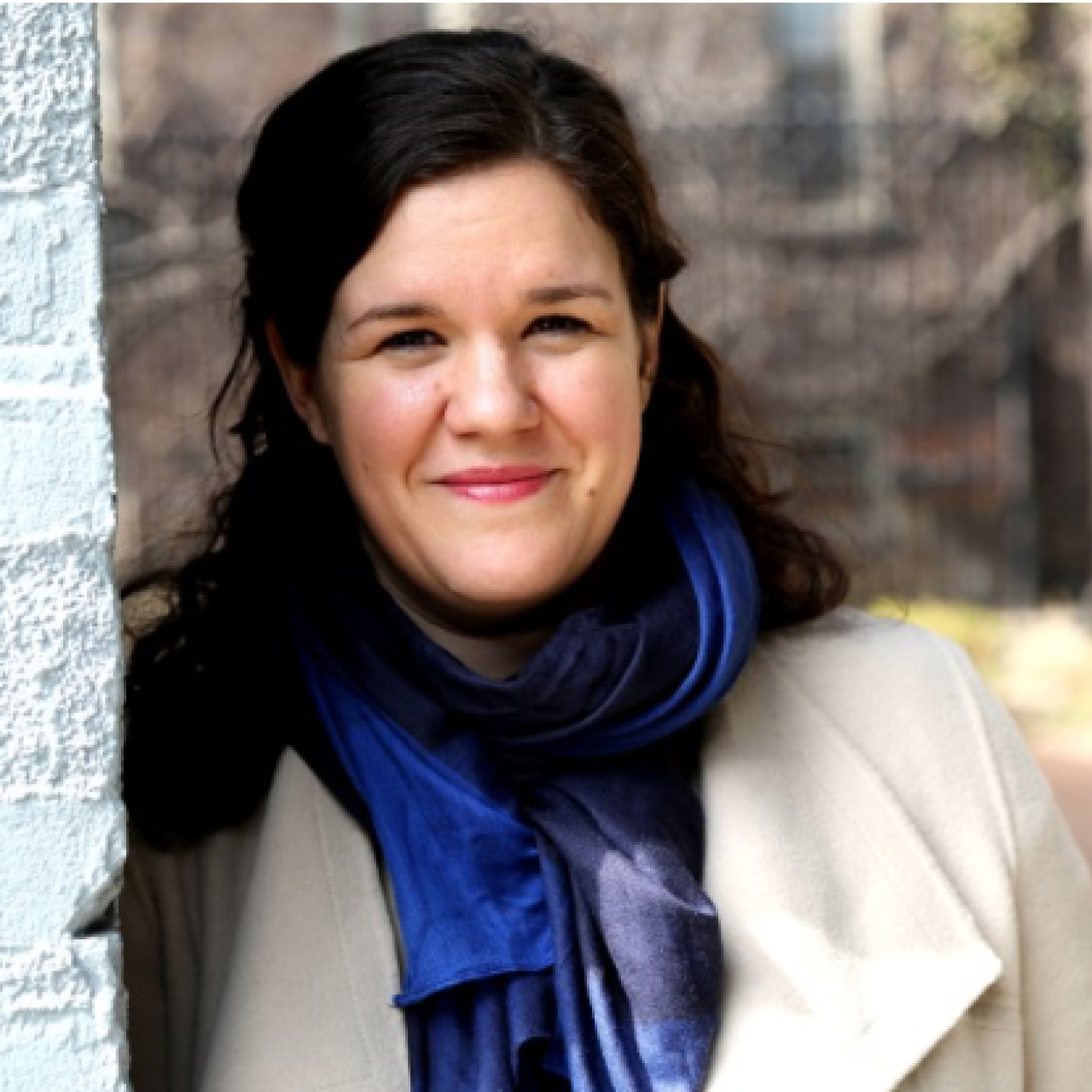 Headshot of Heather Tsavaris in a white coat and blue scar leaning to her left against a white brick wall