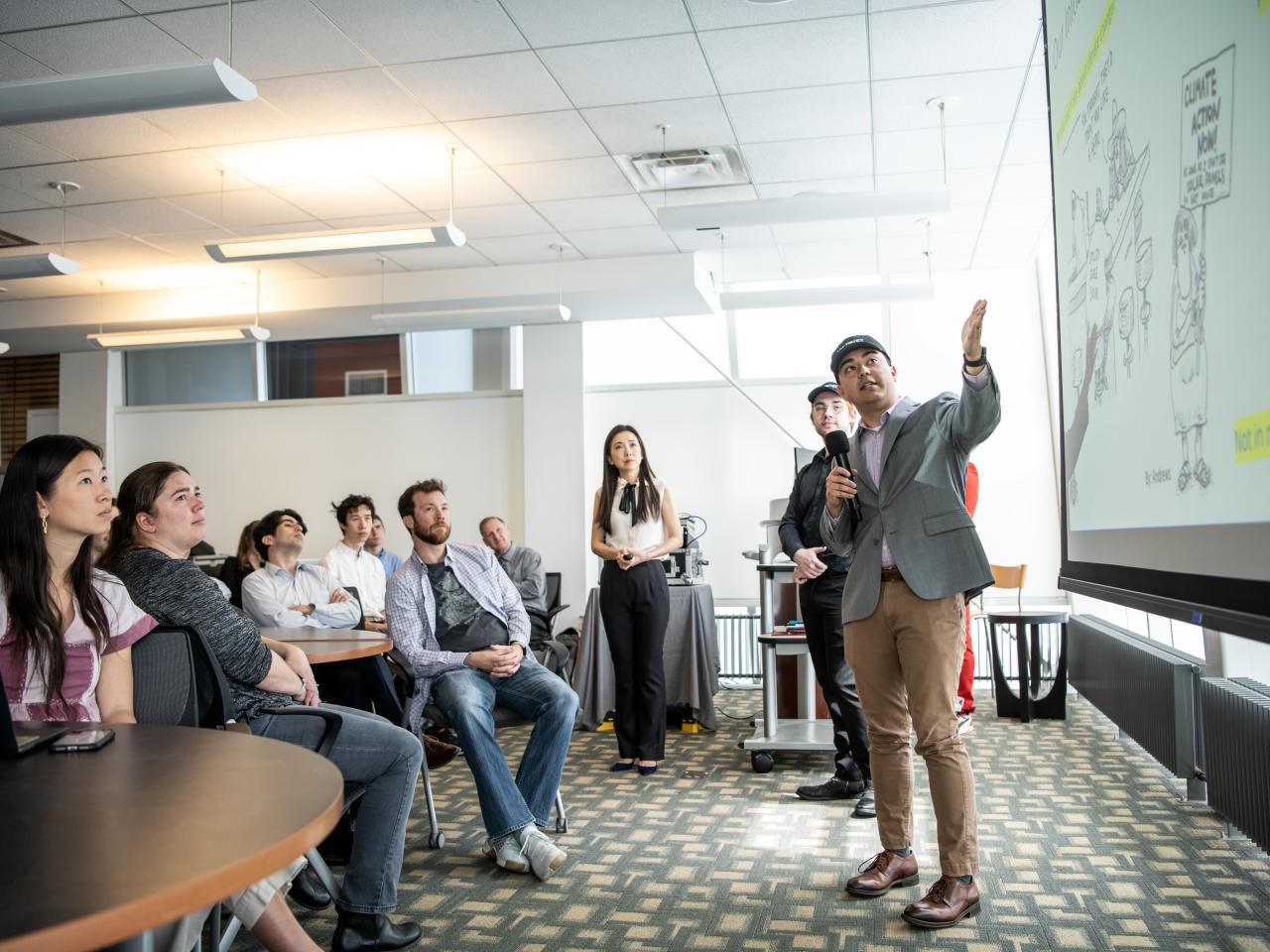 Group of students on the left giving and directing a presentation to an audience on the left.