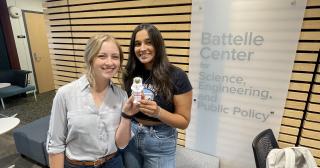 Two students holding a foam astronaut in front of the Battelle Center Logo