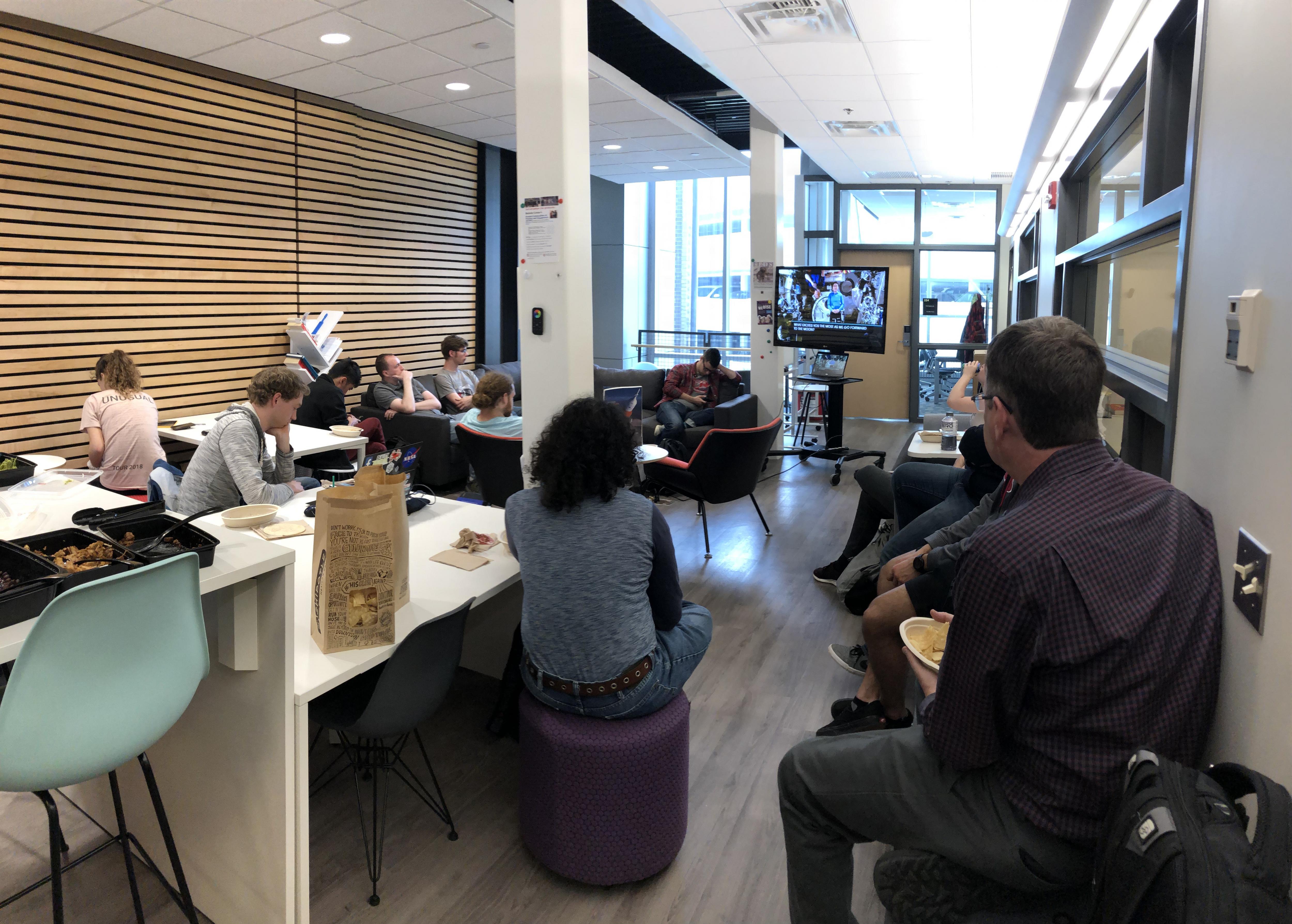 An open space lit in the back. People are gathered and listening to a speaker who is seated and presenting a video at the back of the photograph. 