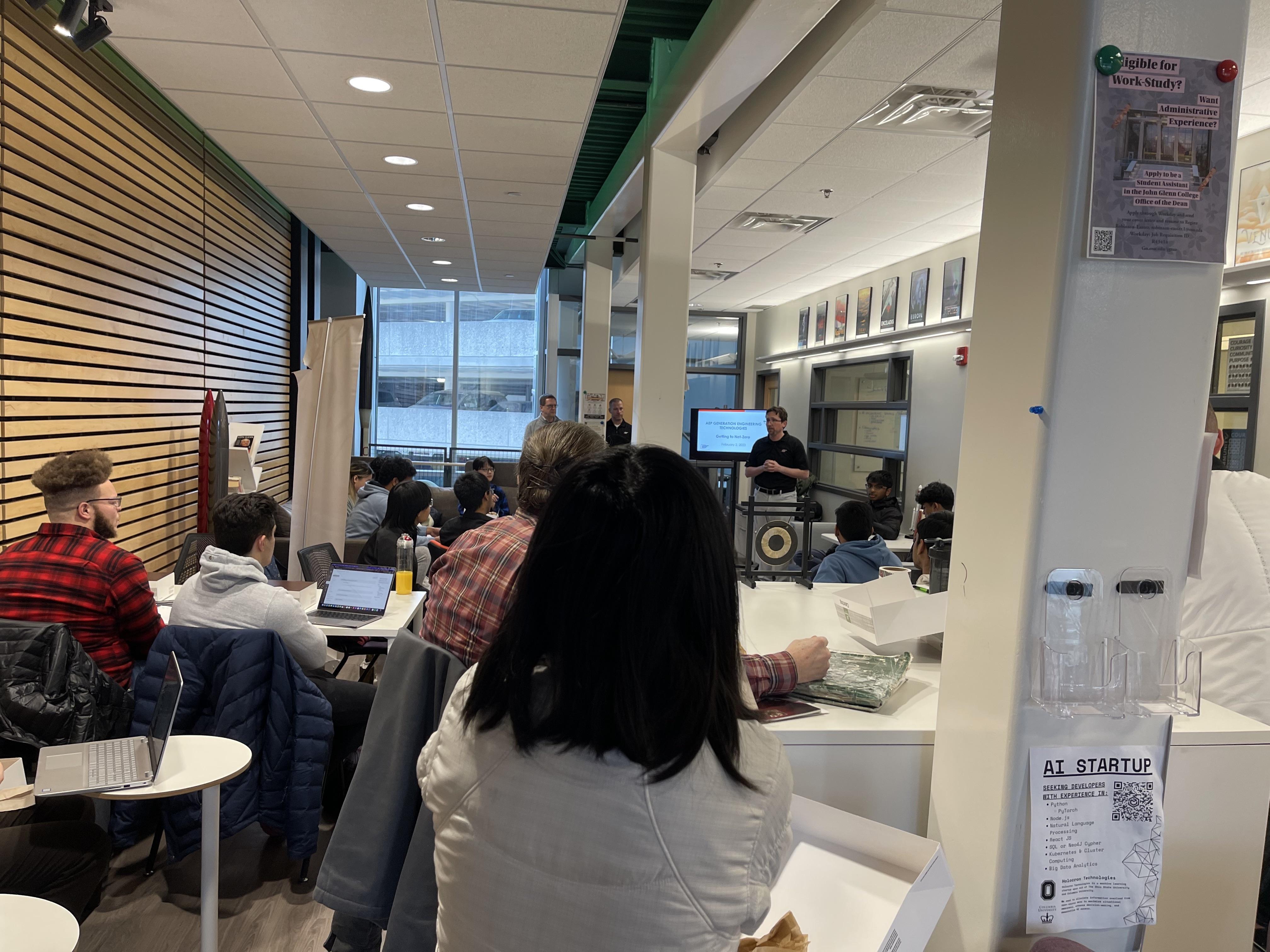 Group of students in a large lounge listening to a presenter at the front with a TV presentation