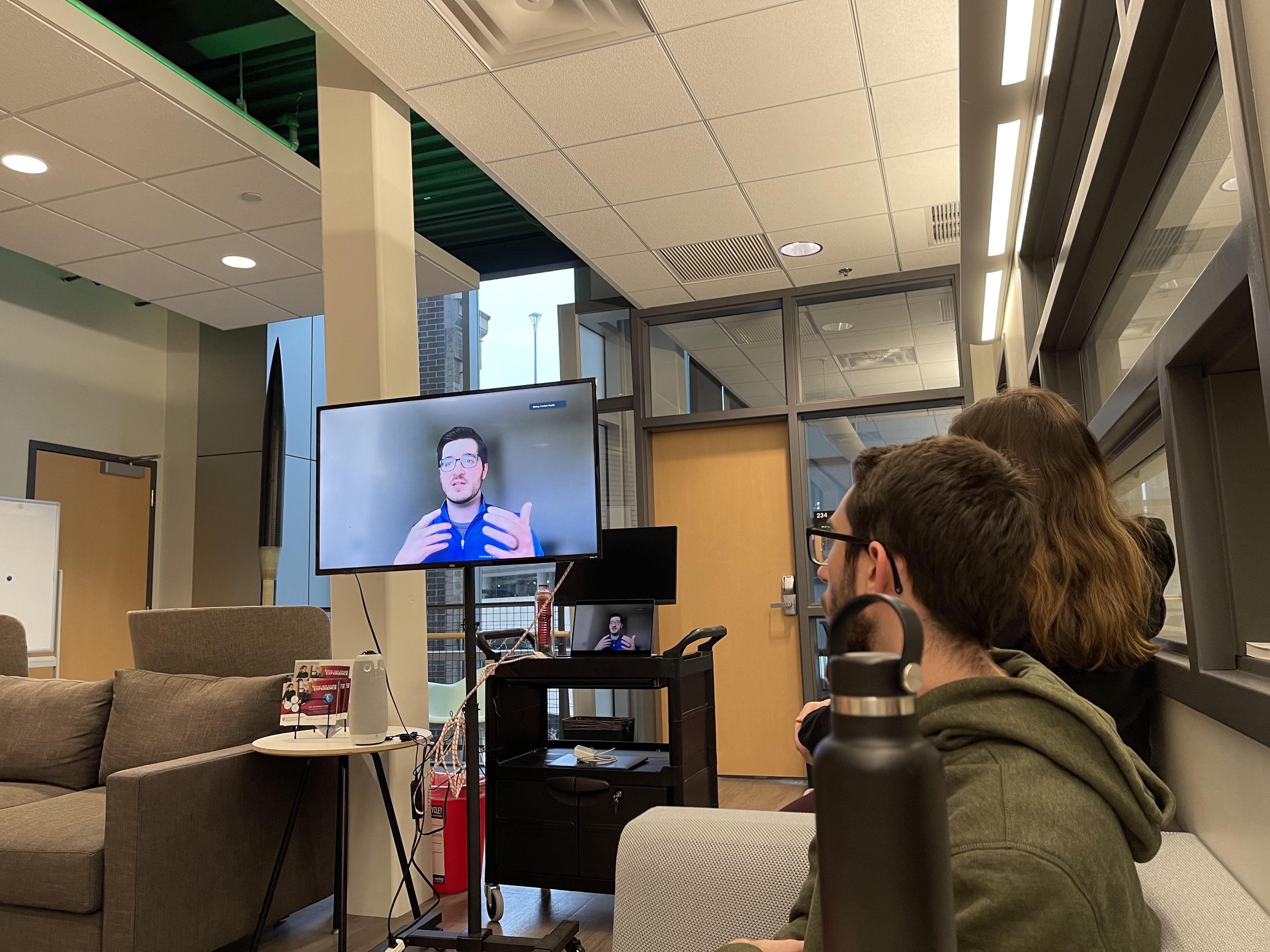 Guest speaker on a monitor speaking with students in a lounge area