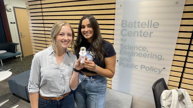 Two students holding a foam astronaut in front of the Battelle Center Logo