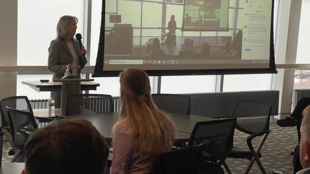 Woman speaking at the front of the room beside a projector screen of the room